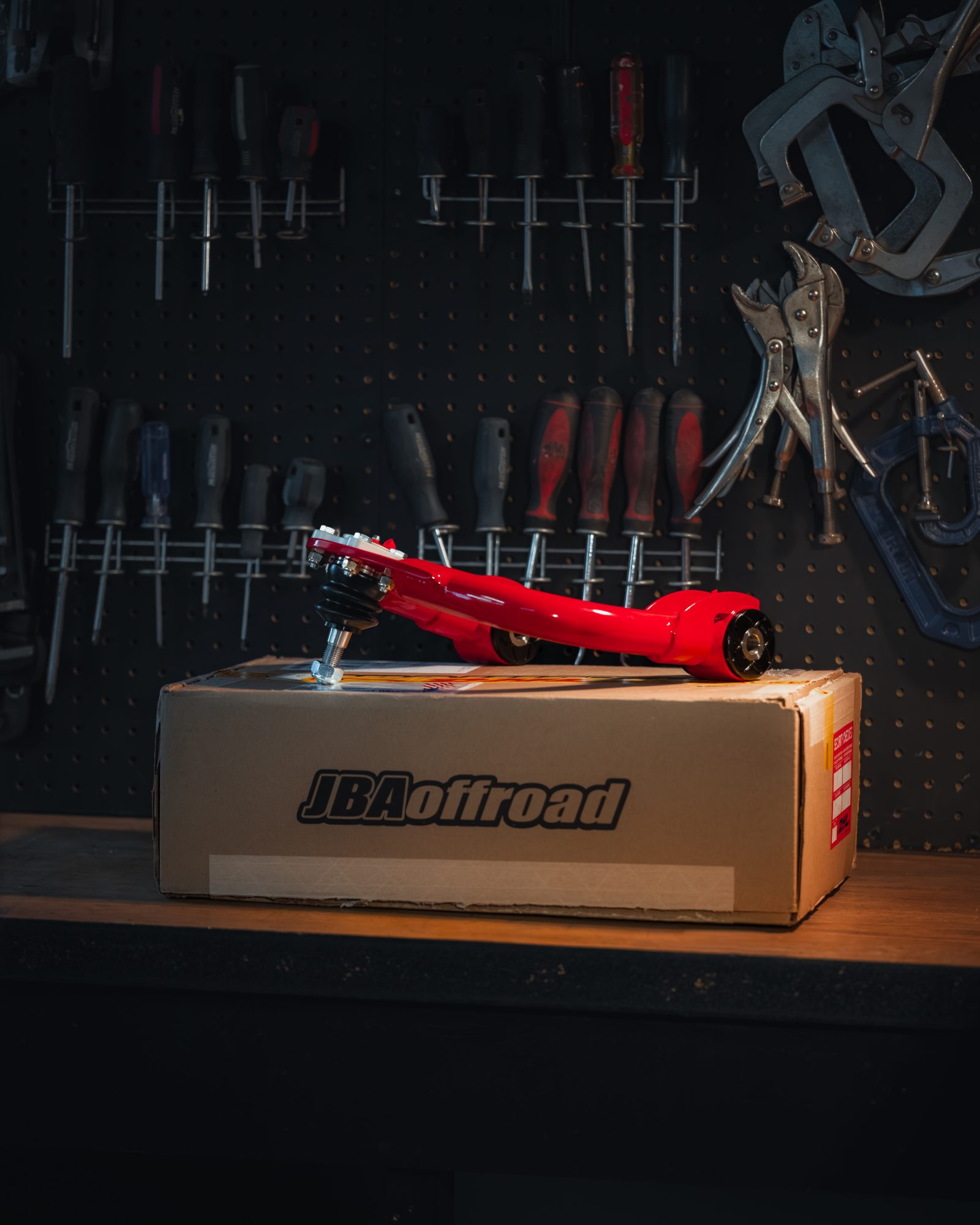 Red JBA STD High Caster upper control arm for the 2nd gen Tacoma on its JBA Offroad box on a workbench